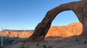 The sun rises behind famous Corona Arch near Moab, Utah. The natural sandstone arch casts a long shadow as the sun illuminates the beautiful red rock formations in the distance. - USA