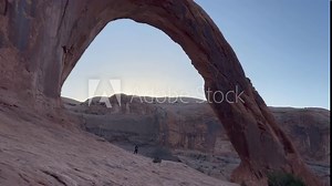 Classic, wide static shot of Corona Arch in Moab, Utah at dawn. A hiker stands underneath the arch to highlight the scale of this massive natural rock formation - USA Stock Video